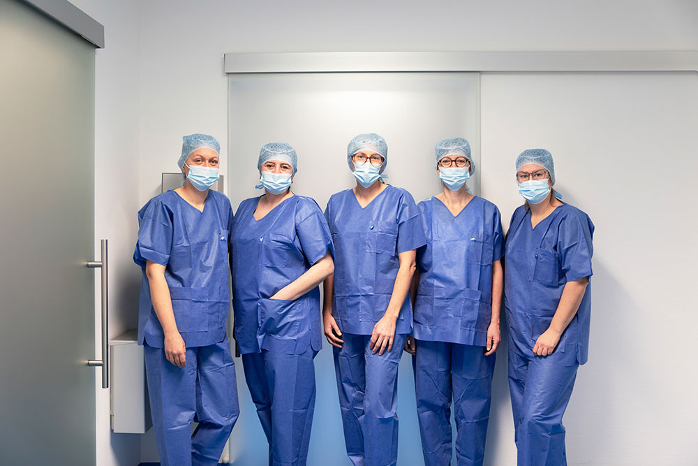 OP-Team vor dem Operationssaal Fünf Mitglieder des OP-Teams in blauen Kitteln und Masken vor dem Operationssaal.
